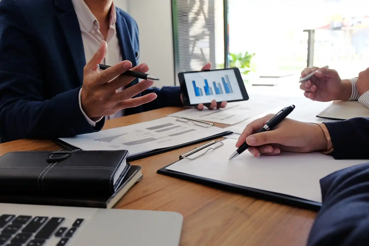 Businessmen are seated at a table. One is holding a small tablet with charts, presenting them to 2 people sat across the table, both taking notes