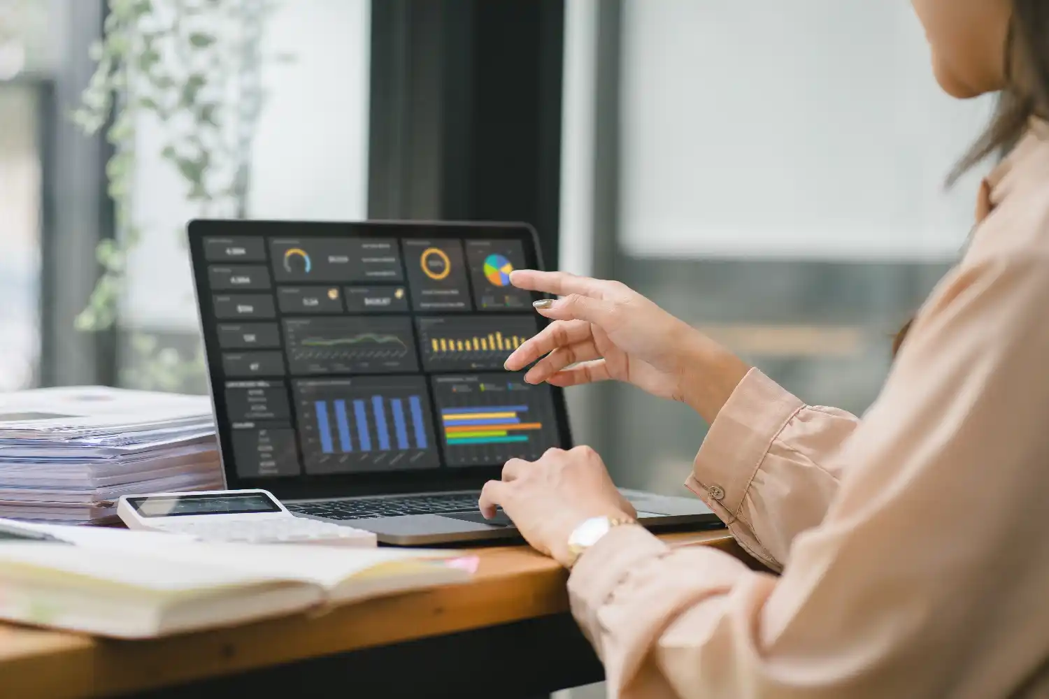 A woman seated at a desk has a log book open with a calculator sitting on it. She is analyzing a dashboard displaying some financial data